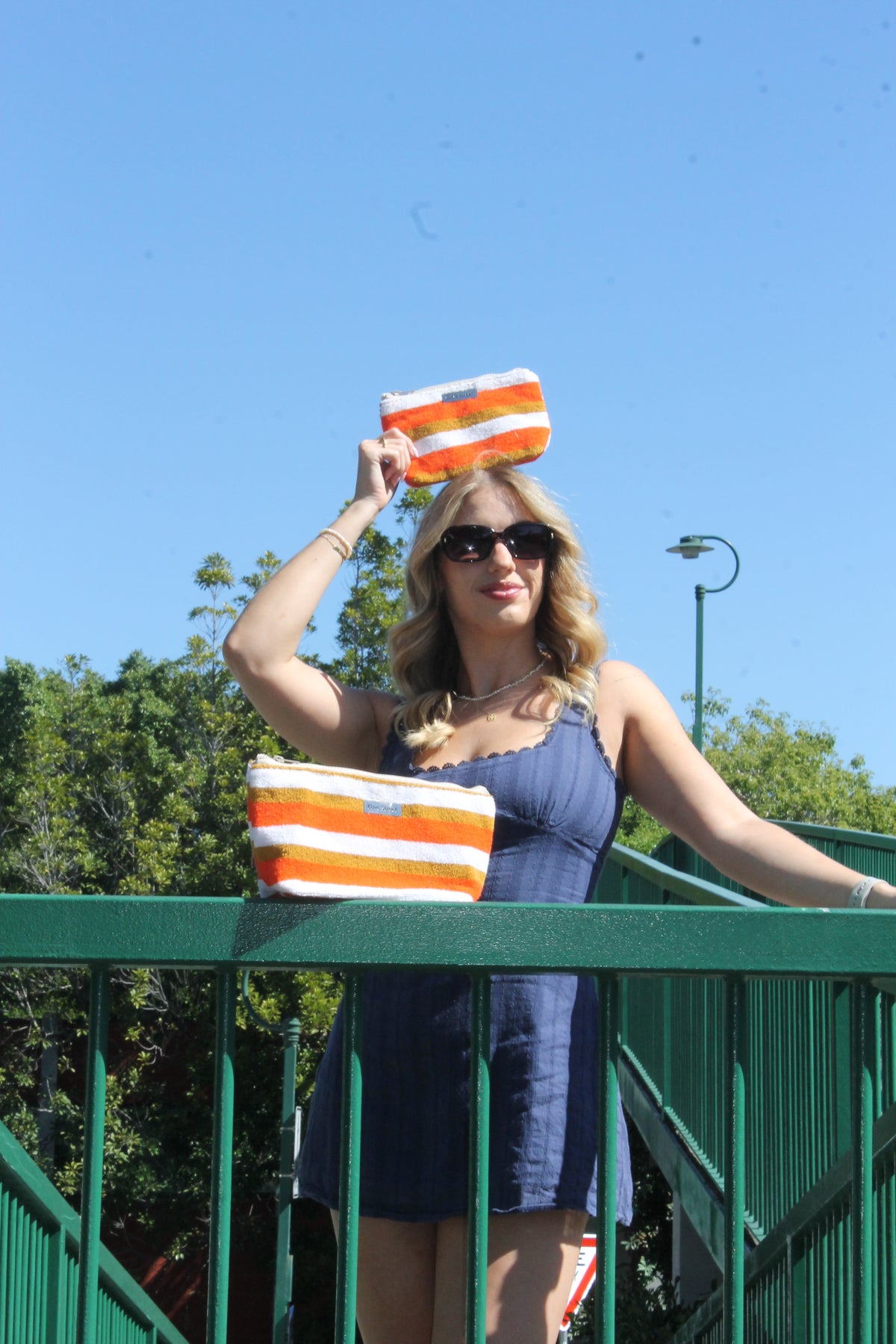 A woman wearing a blue dress and sunglasses stands outdoors on a green bridge, holding a striped orange, white, and blue pouch above her head while another matching pouch rests on the railing.